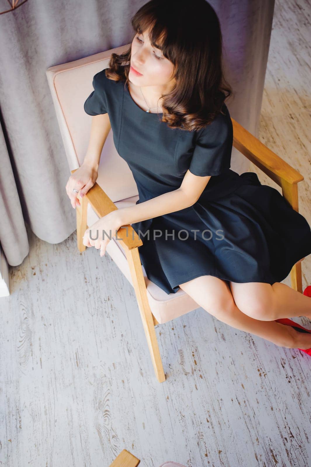 Young attractive woman with long legs in black elegant dress, sits in chair near window in interior of room.