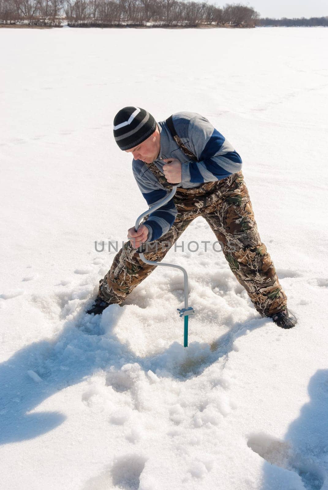 Winter fishing on the river. Young man drill a hole in the ice.