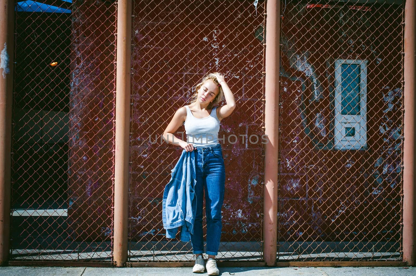 young woman in jeans clothes outdoors. portrait of a girl with freckles on her face, stylish girl on a background of a fence of a grid, on a sunny summer day. Holds a denim jacket in his hand