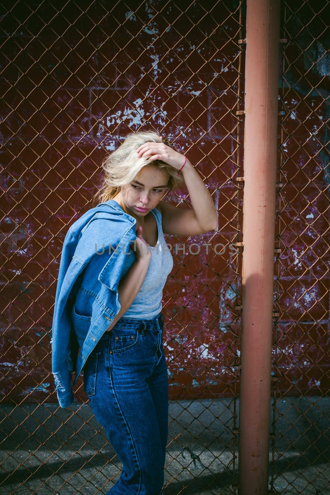 young woman in jeans clothes outdoors. portrait of a girl with freckles on her face, stylish girl on a background of a fence of a grid, on a sunny summer day. Holds a denim jacket in his hand