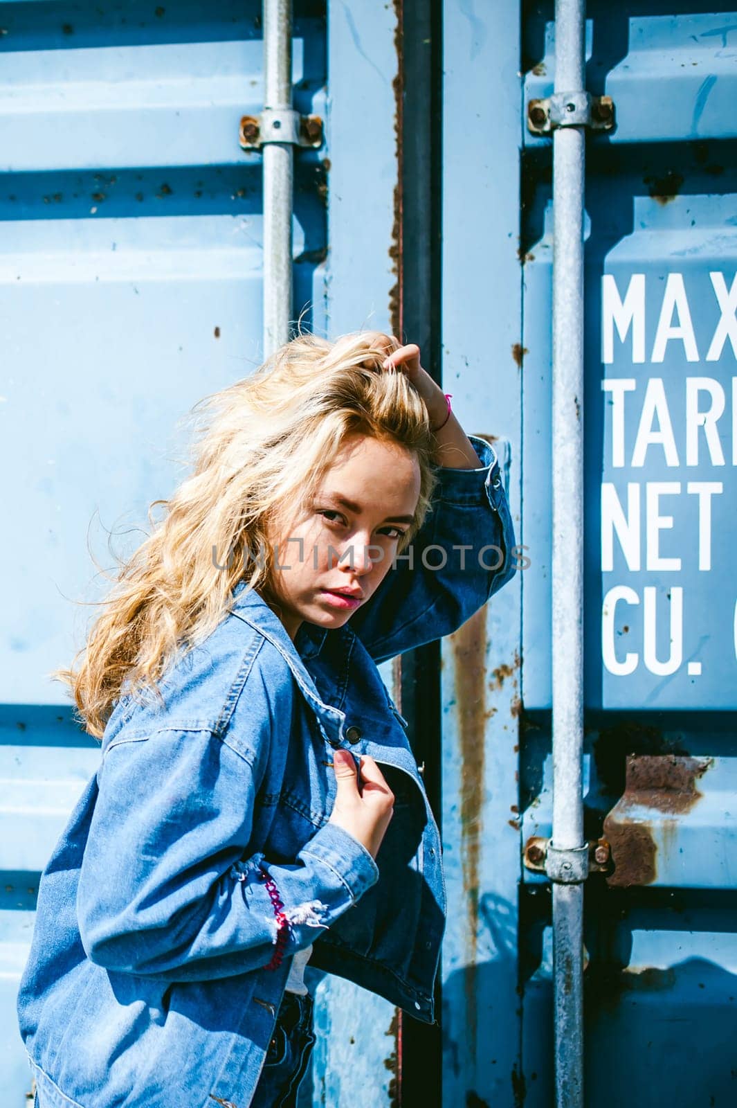 young beautiful woman in jeans clothes outdoors. portrait of a girl with freckles on her face, stylish girl on a freight containers background, on a sunny summer day. wind develops her hair