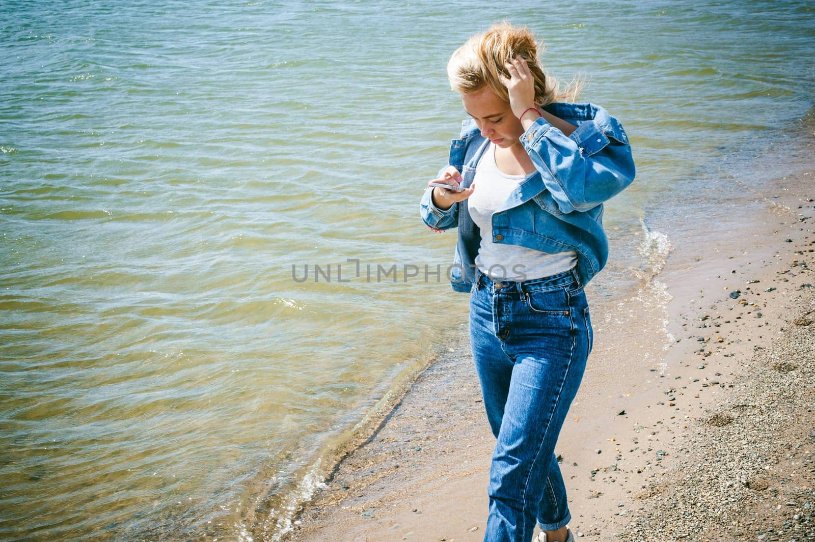 young beautiful woman in jeans clothes outdoors. portrait of a girl with freckles on her face, stylish girl on sea beach, on a sunny summer autumn day. looks at mobile phone