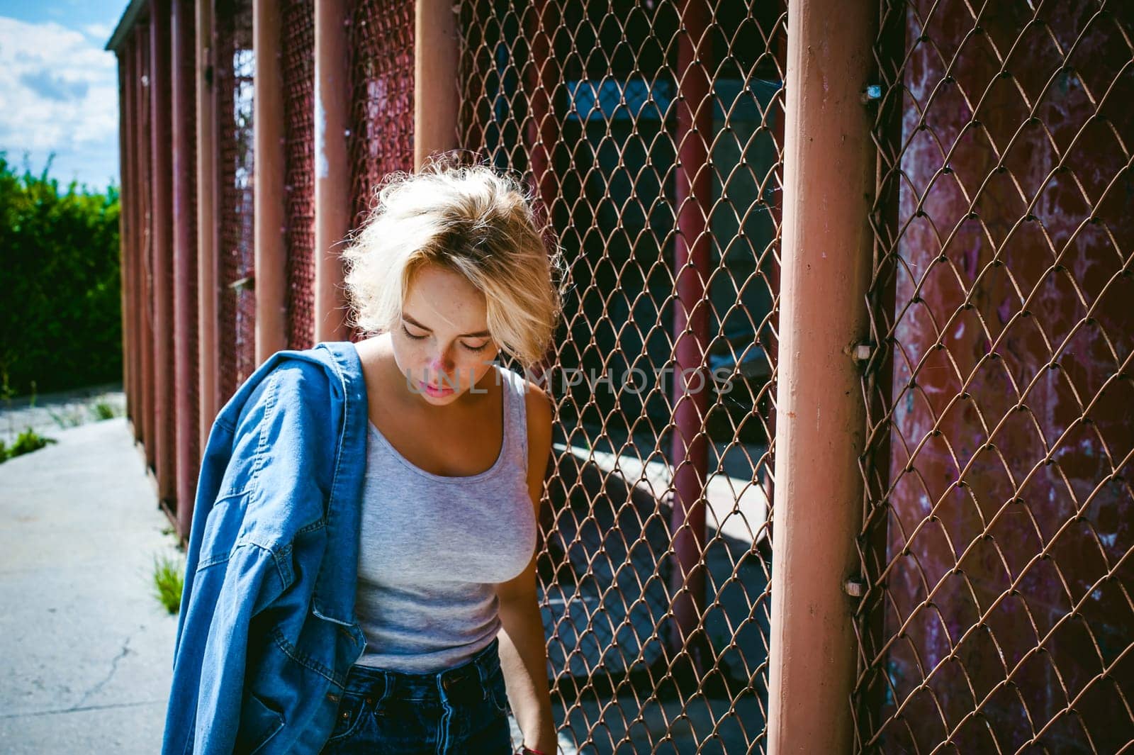 young woman in jeans clothes outdoors. portrait of a girl with freckles on her face, stylish girl on a fence of a grid, a sunny summer day...