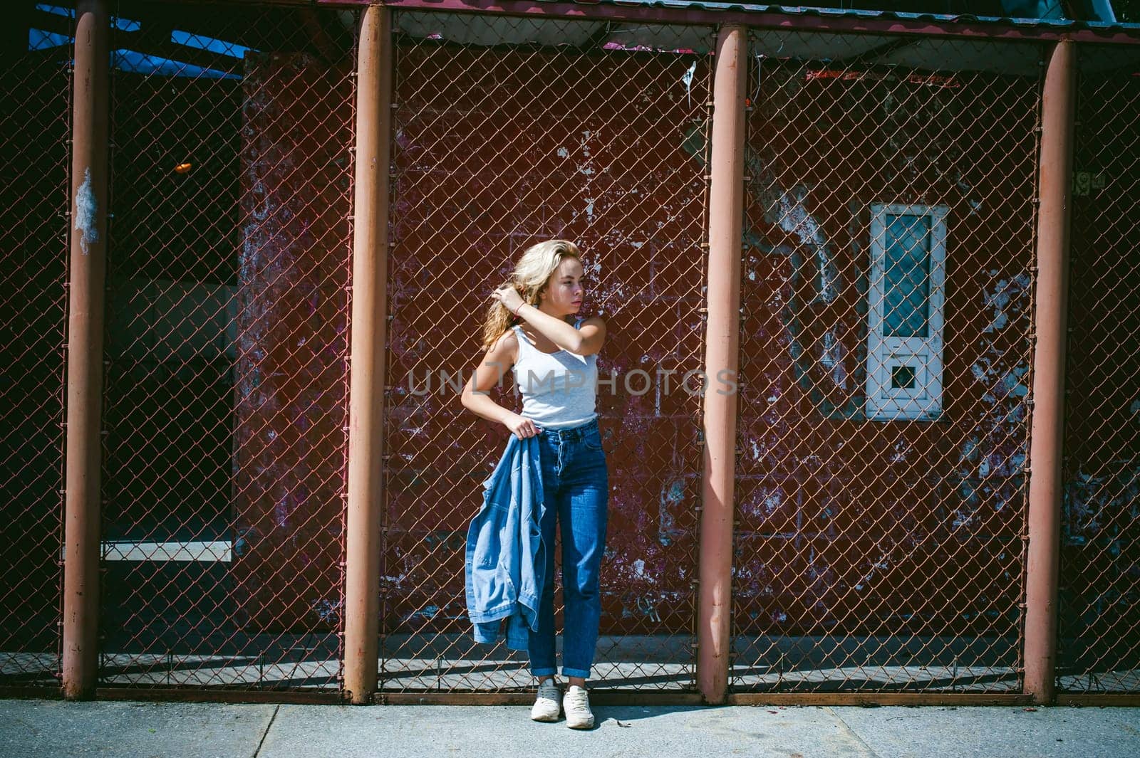young woman in jeans clothes outdoors. portrait of a girl with freckles on her face, stylish girl on a background of a fence of a grid, on a sunny summer day. Holds a denim jacket in his hand