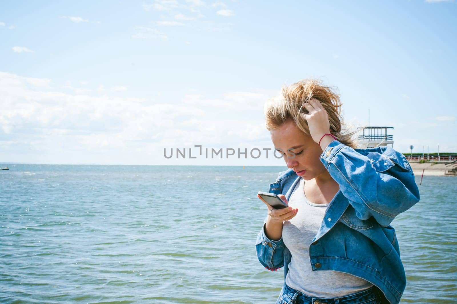 young beautiful woman in jeans clothes outdoors. portrait of a girl with freckles on her face, stylish girl on sea beach, on a sunny summer autumn day. looks at mobile phone