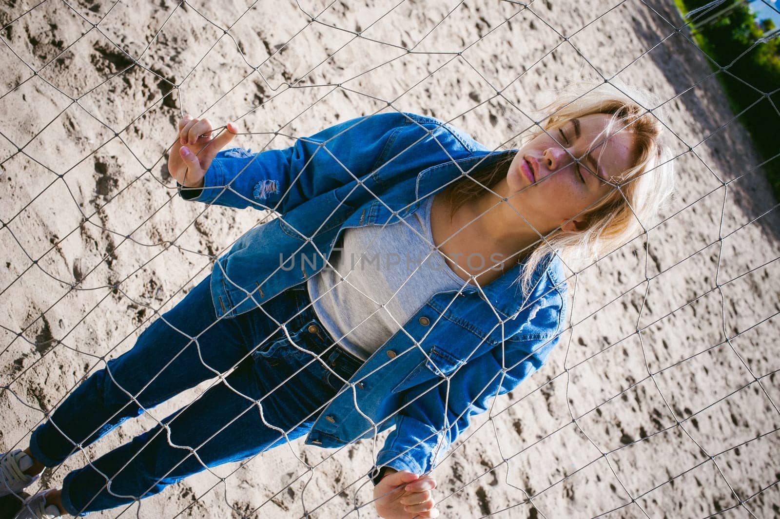 young woman in jeans clothes outdoors. portrait of a girl with freckles on her face, against the background of a football gate grid, on a sunny summer day