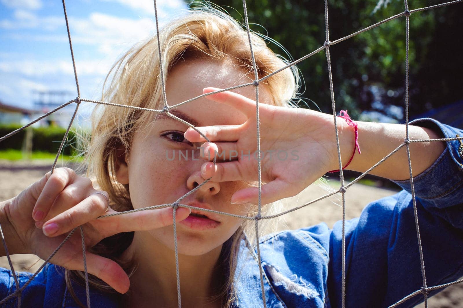 young woman in jeans clothes outdoors. portrait of a girl with freckles on her face, against the background of a football gate grid, on a sunny summer day. hands and fingers