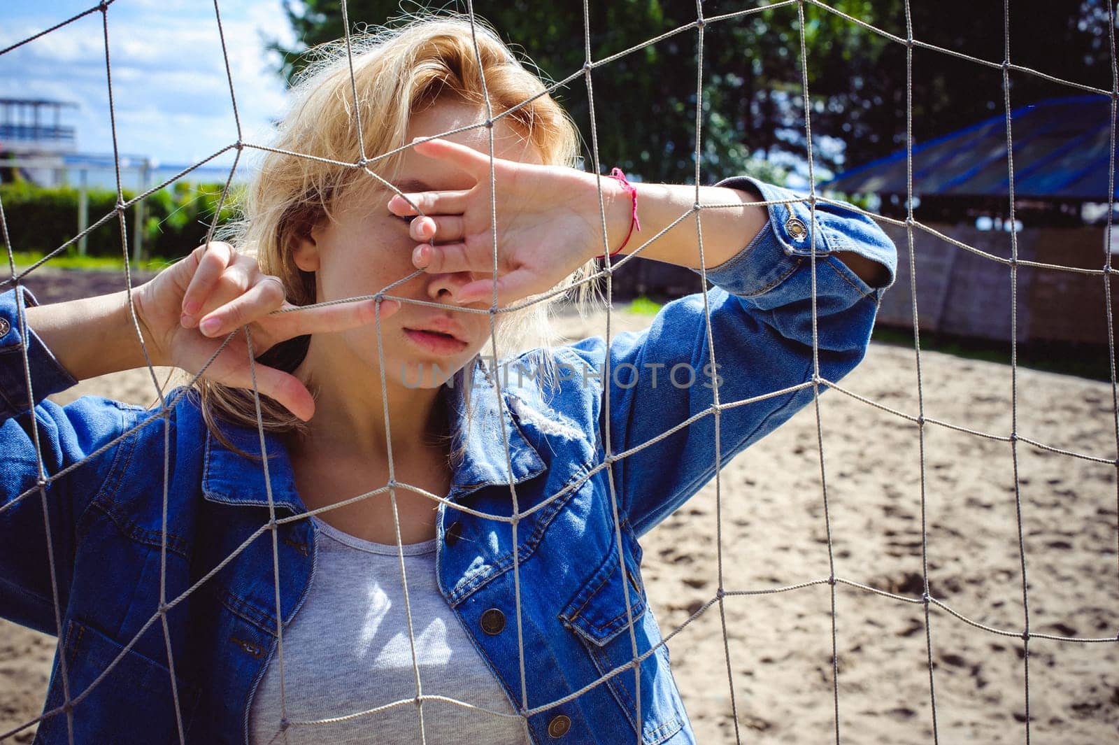 young woman in jeans clothes outdoors. portrait of a girl with freckles on her face, against the background of a football gate grid, on a sunny summer day. hands and fingers