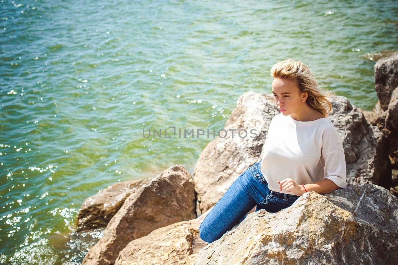 Portrait young woman on stones near sea. Beautiful girl is resting on coast, enjoying outdoor recreation.