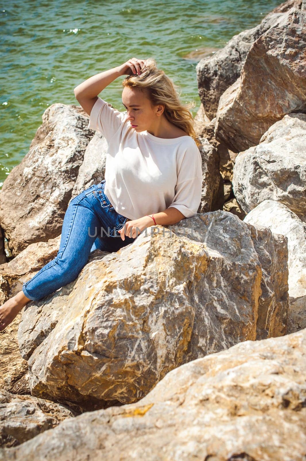Portrait young woman on stones near sea. Beautiful girl is resting on coast, enjoying outdoor recreation.