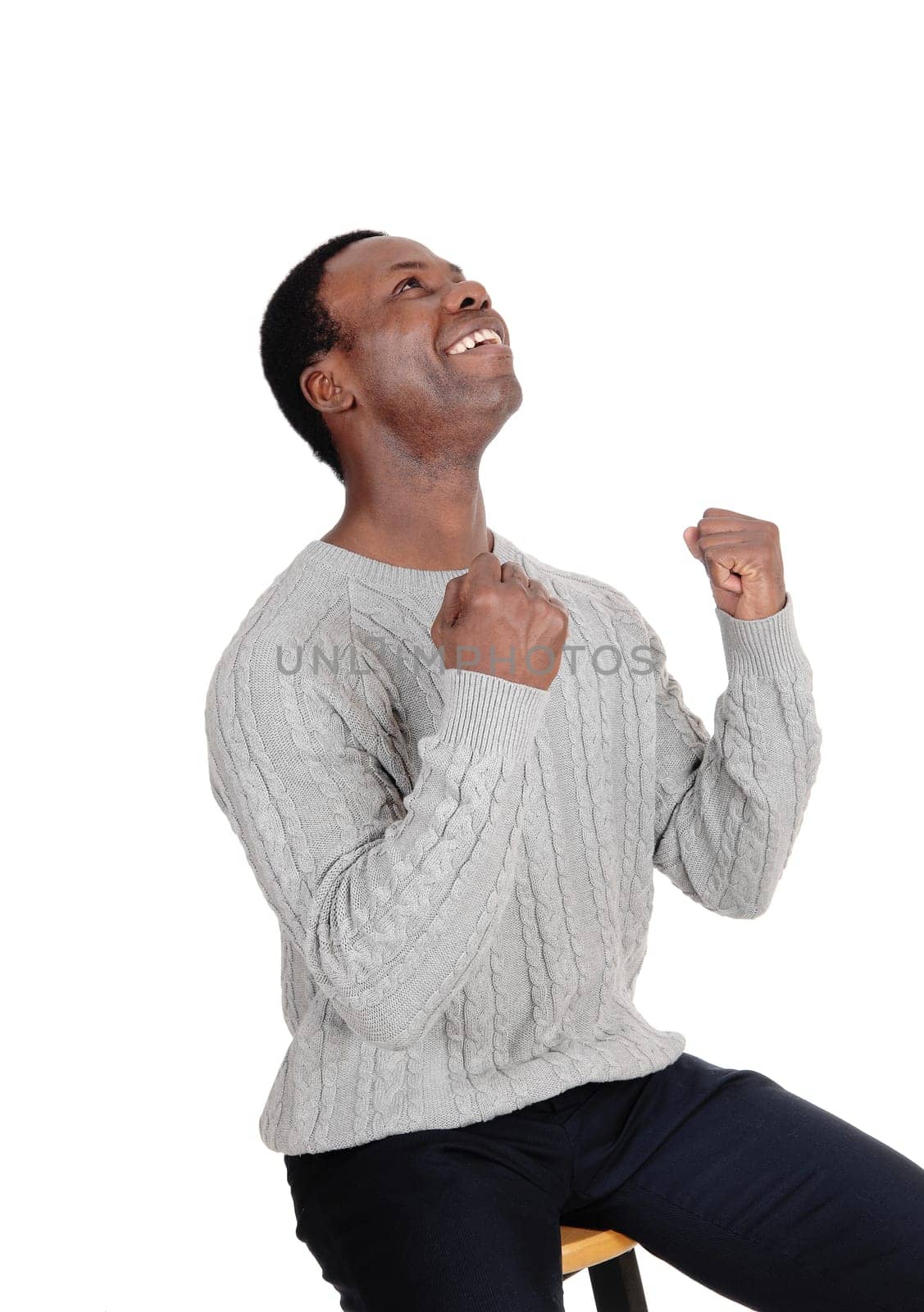 A young handsome African American man sitting in a sweater with.his fists and smiling of victory and success, isolated over white.