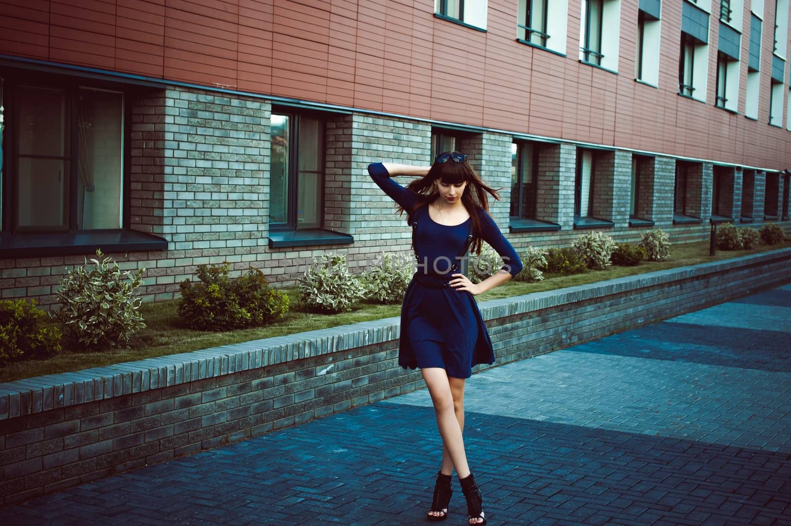 Young attractive woman with long legs with long hair in a blue elegant dress with a sword belt, strolls in a European modern courtyard, a warm summer day