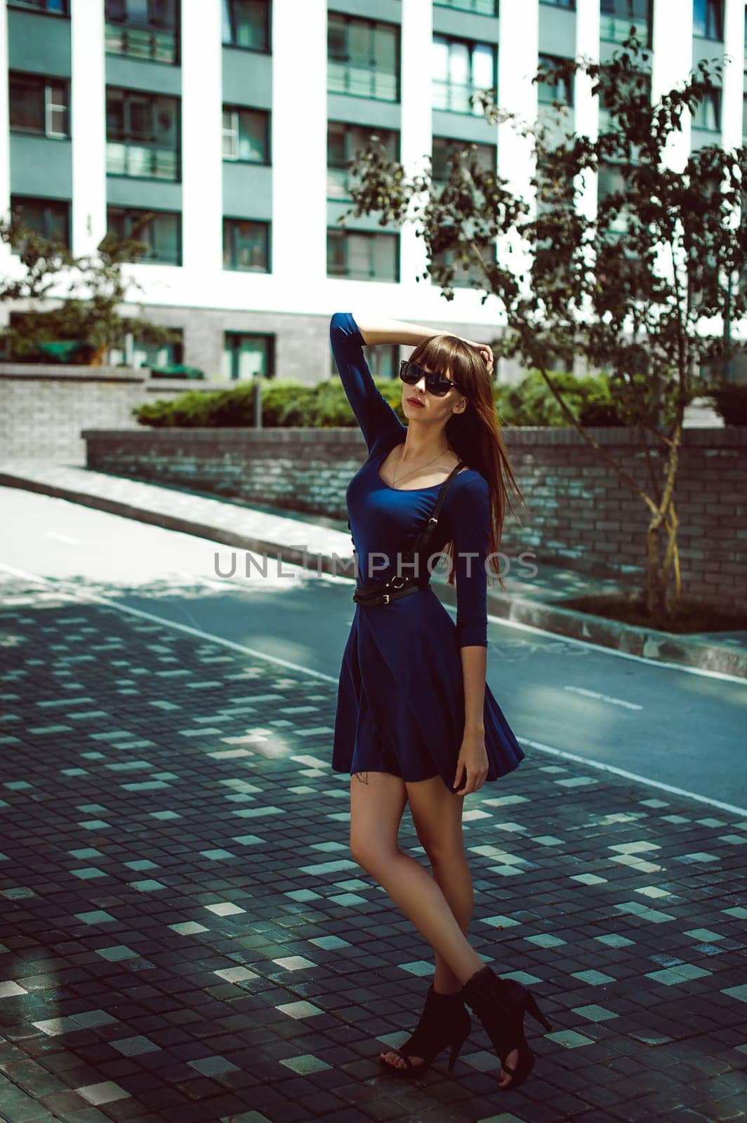 Young attractive woman with long legs with long hair in a blue elegant dress with a sword belt In sunglasses, strolls in a European modern courtyard, a warm summer day