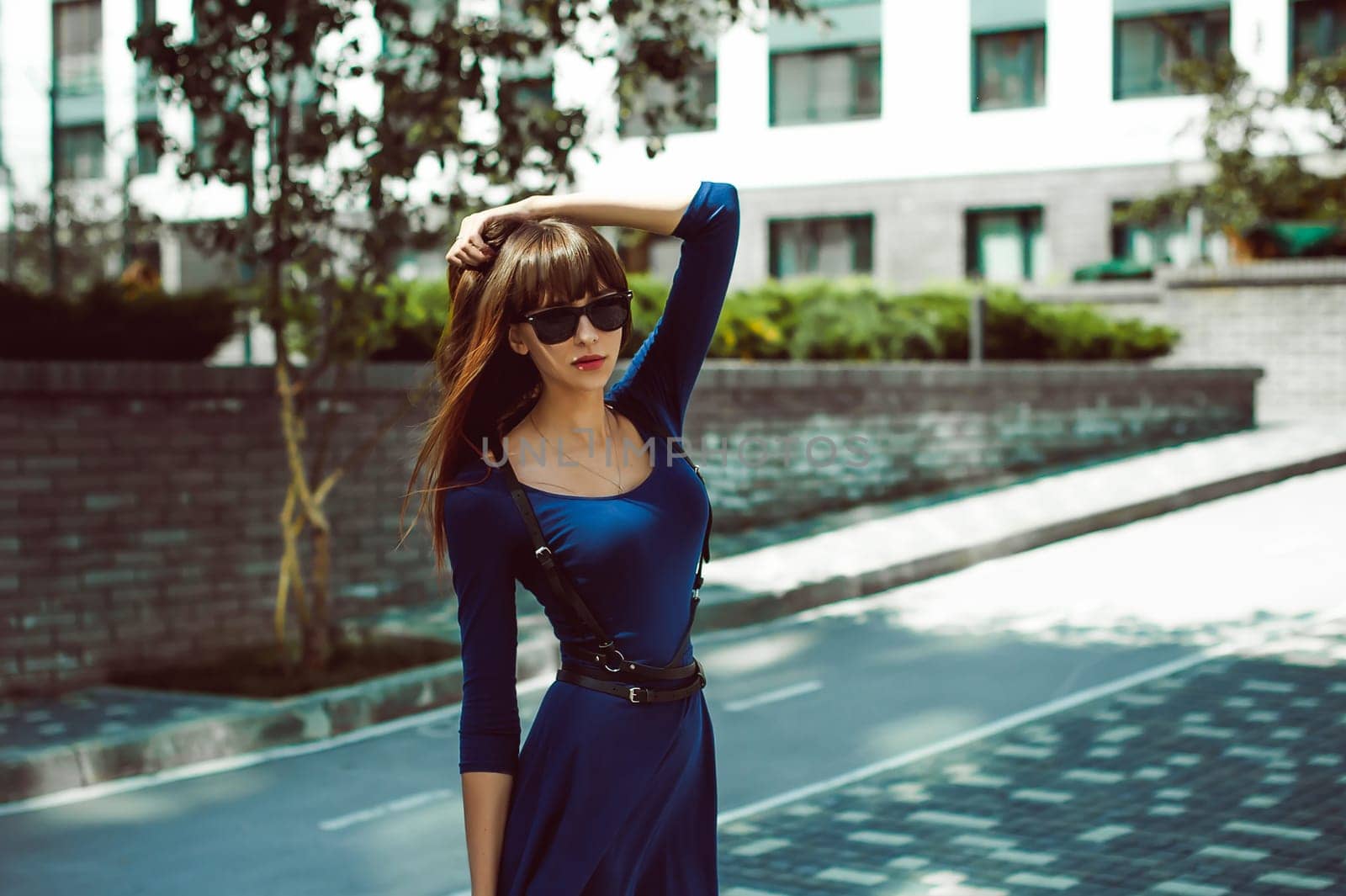 Young attractive woman with long legs with long hair in a blue elegant dress with a sword belt In sunglasses, strolls in a European modern courtyard, a warm summer day