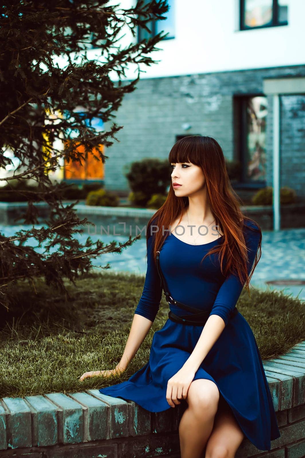 Young attractive woman with long legs with long hair in a blue elegant dress with a sword belt, strolls in a European modern courtyard, a warm summer day