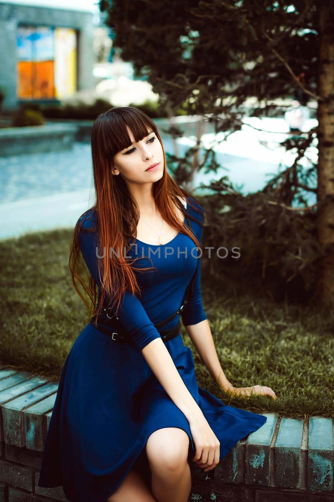 Young attractive woman with long legs with long hair in a blue elegant dress with a sword belt, strolls in a European modern courtyard, a warm summer day