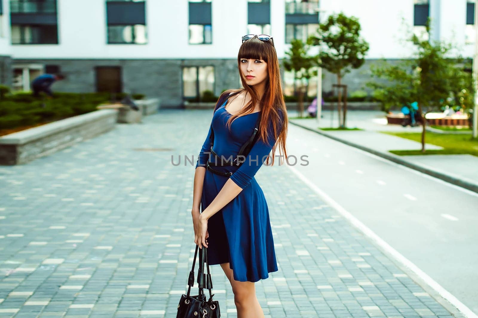 Young attractive woman with long legs with long hair in a blue elegant dress with a sword belt with a black bag in his hand, strolls in a European modern courtyard, a warm summer day