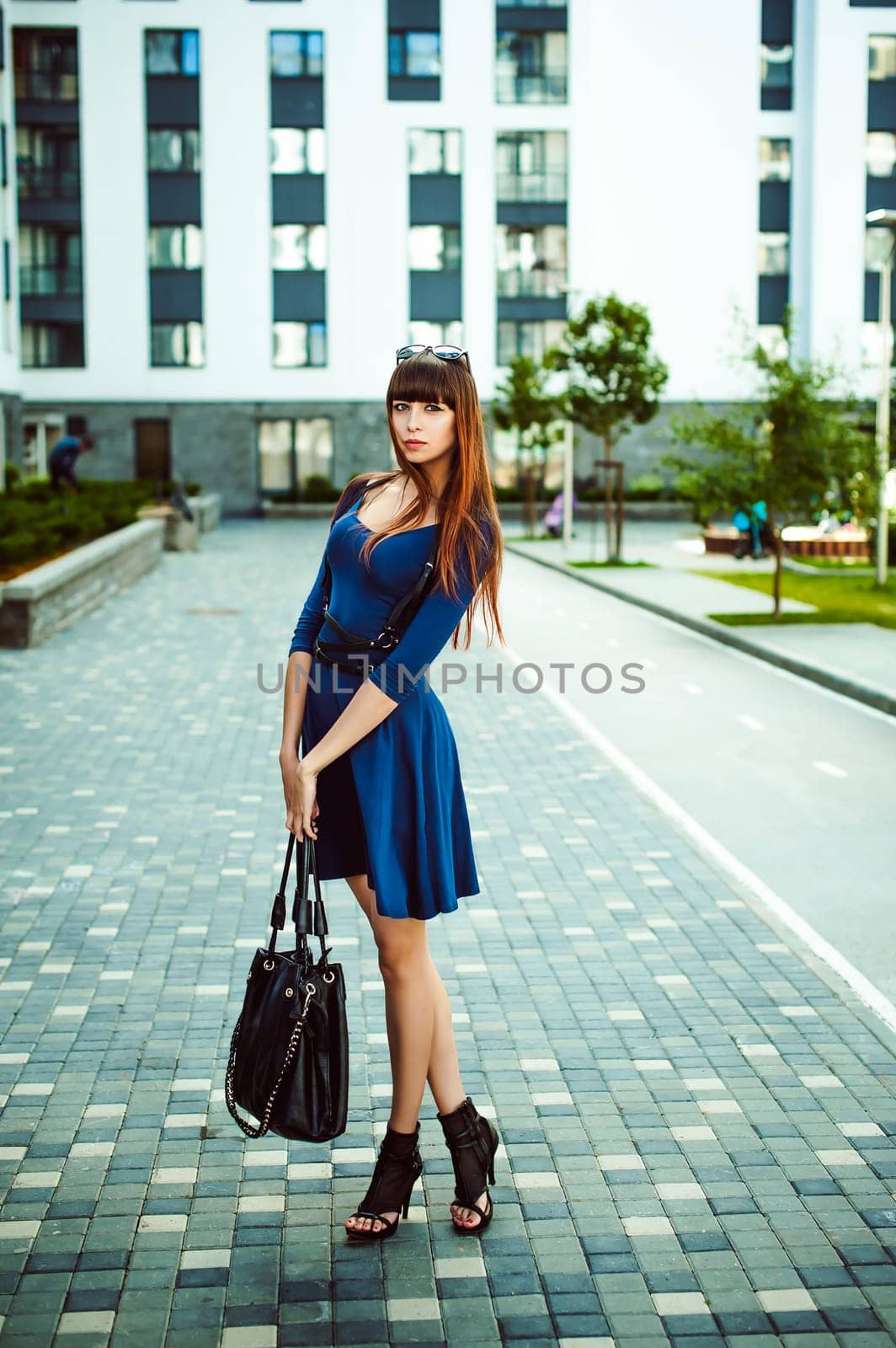 Young attractive woman with long legs with long hair in a blue elegant dress with a sword belt with a black bag in his hand, strolls in a European modern courtyard, a warm summer day