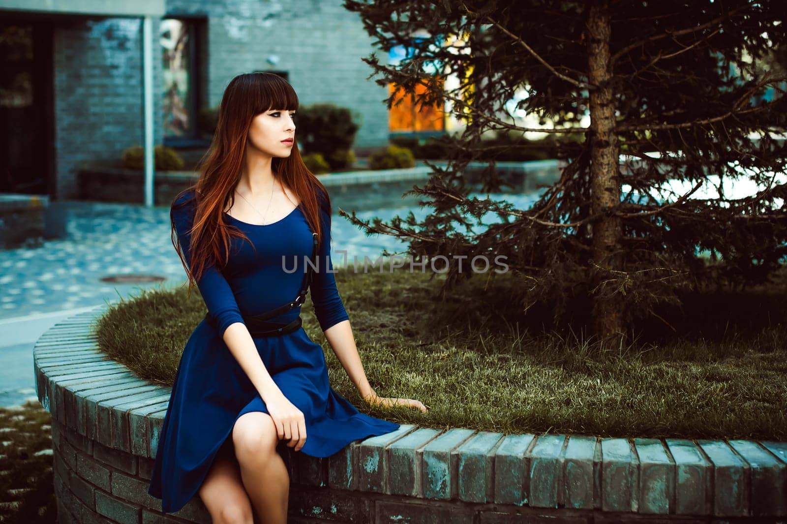 Young attractive woman with long legs with long hair in a blue elegant dress with a sword belt, strolls in a European modern courtyard, a warm summer day