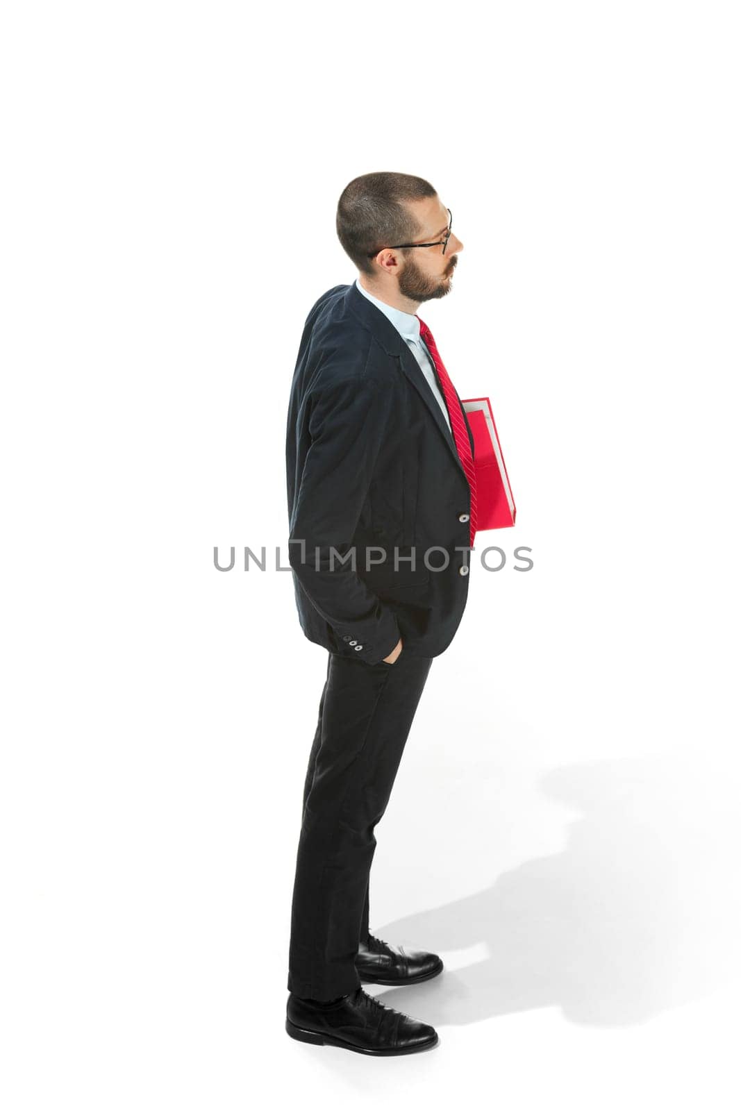 Full body or full-length profile of businessman with red folder on white studio background. Serious bearded young man in glasses, suit, red tie standing in office. Business, career, success concept.