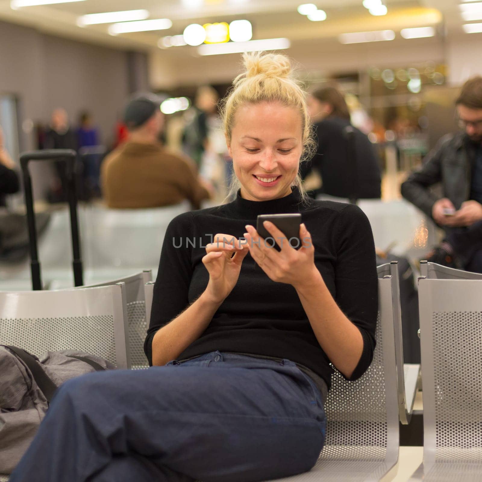 Casual blond young woman using her mobile phone while waiting to board a plane at airport departure gates.
