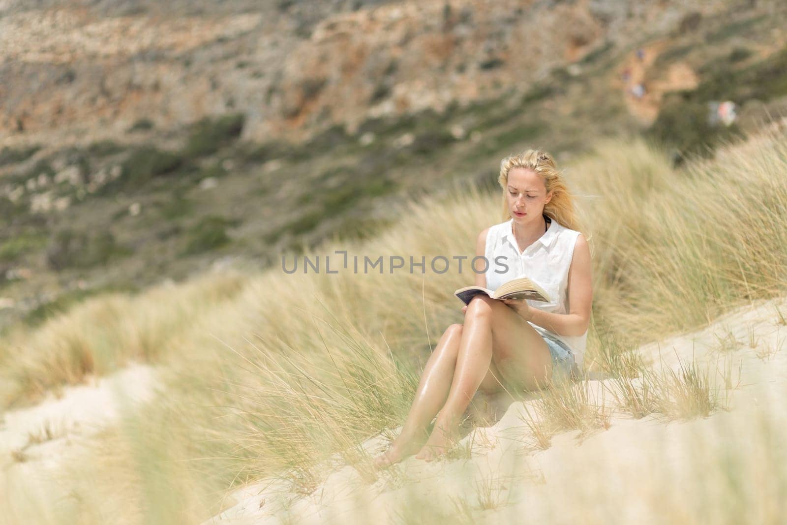 Relaxed woman enjoying sun, freedom and good book an beautiful sandy beach of Balos in Greece. Young lady reading, feeling free and relaxed. Vacations, freedom, happiness, enjoyment and well being.