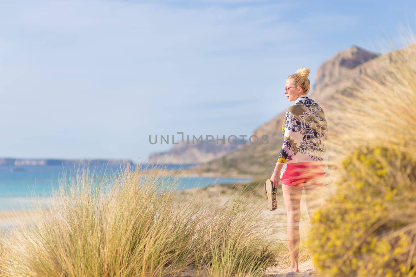 Relaxed woman wrapped in colorful scarf, enjoying sun, freedom and life at beautiful Balos beach in Greece. Concept of holidays, vacations, freedom, joy and well being.