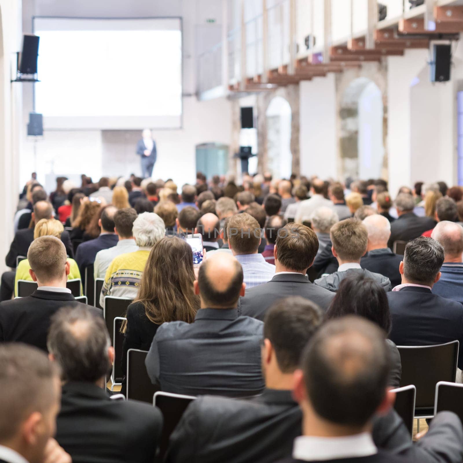 Speaker giving a talk in conference hall at business event. Audience at the conference hall. Business and Entrepreneurship concept. Focus on unrecognizable businessman in the audience.
