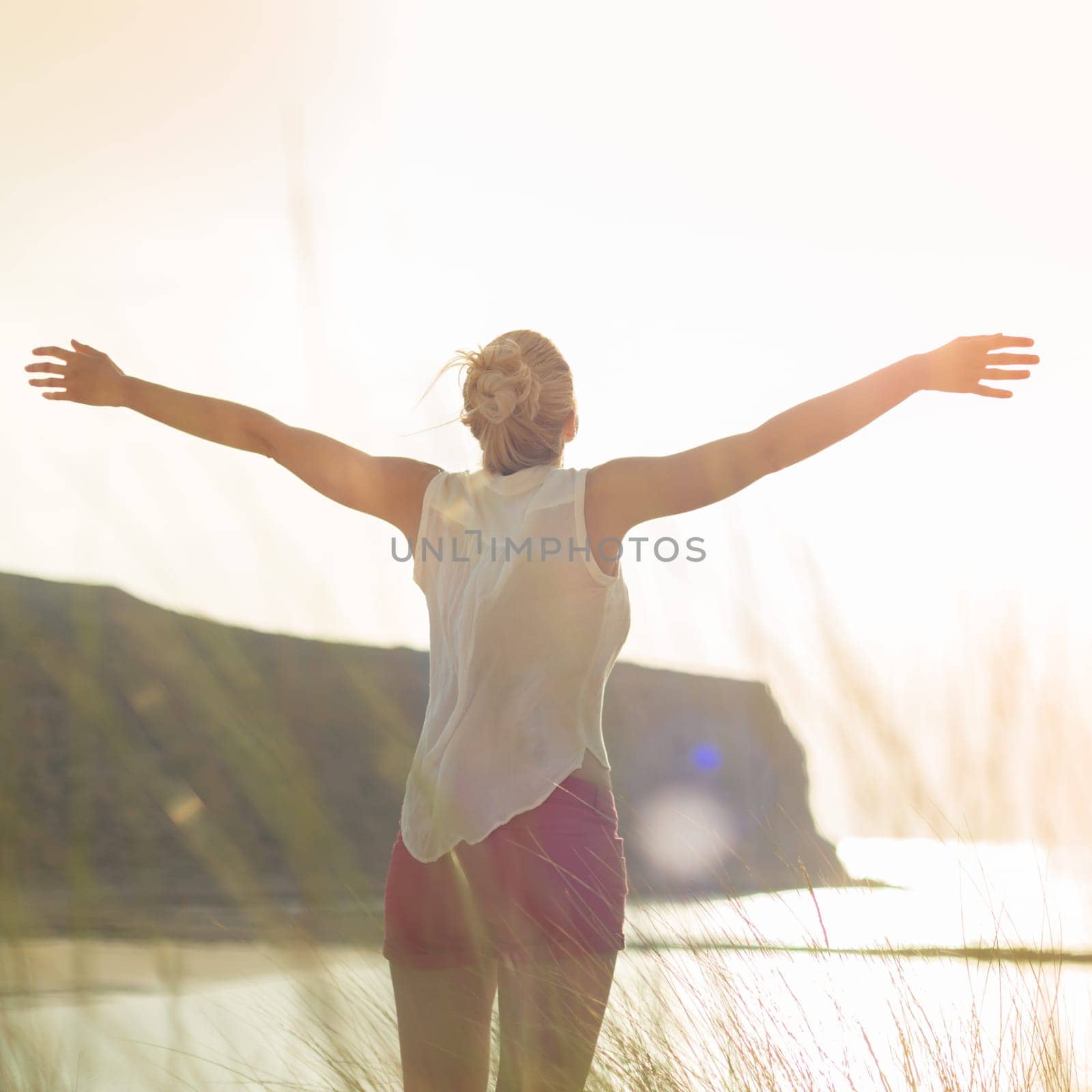 Relaxed woman in white shirt, arms raised, enjoying sun, freedom and life an beautiful beach. Young lady feeling free, relaxed and happy. Concept of vacations, freedom, happiness, joy and well being.