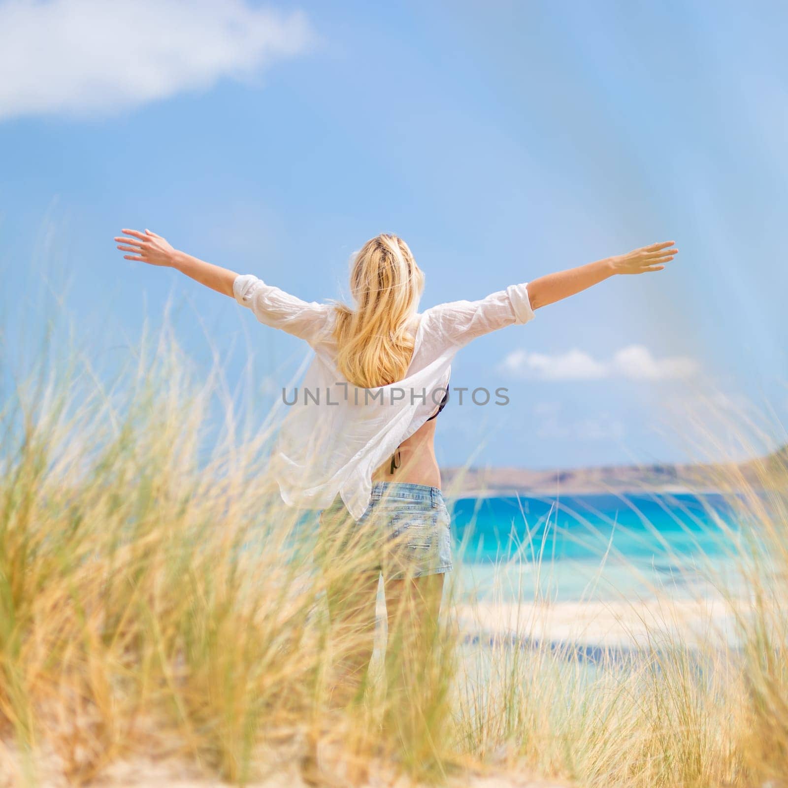 Relaxed woman, arms raised, enjoying sun, freedom and life an a beautiful beach. Young lady feeling free, relaxed and happy. Concept of vacations, freedom, happiness, enjoyment and well being.