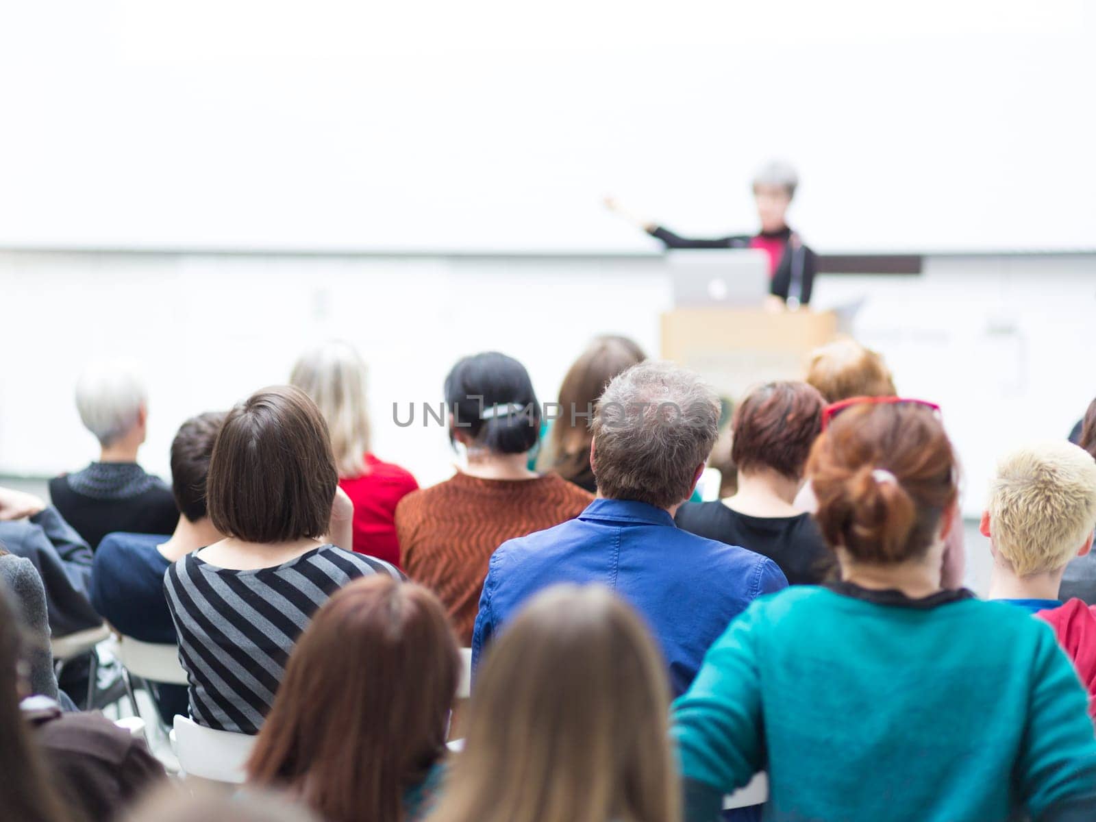 Business and entrepreneurship symposium. Female speaker giving a talk at business meeting. Audience in conference hall. Rear view of unrecognized participant in audience. Copy space on whitescreen.