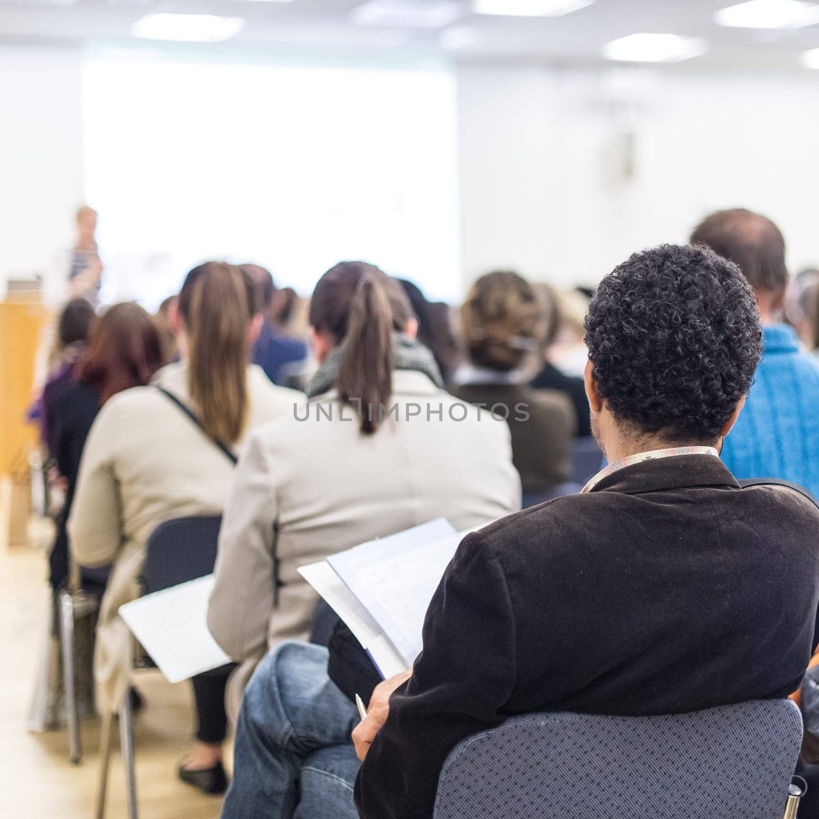 Business and entrepreneurship symposium. Female speaker giving a talk at business meeting. Audience in conference hall. Rear view of unrecognized participants making notes in audience.