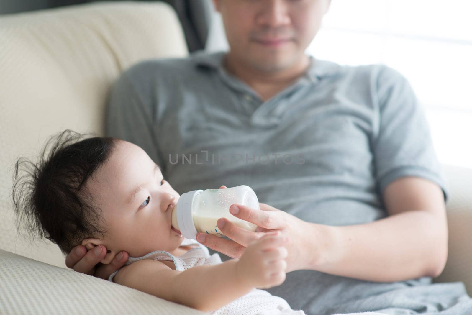 Happy Asian family at home. Father bottle feed formula milk to 9 months old child, living lifestyle indoors.