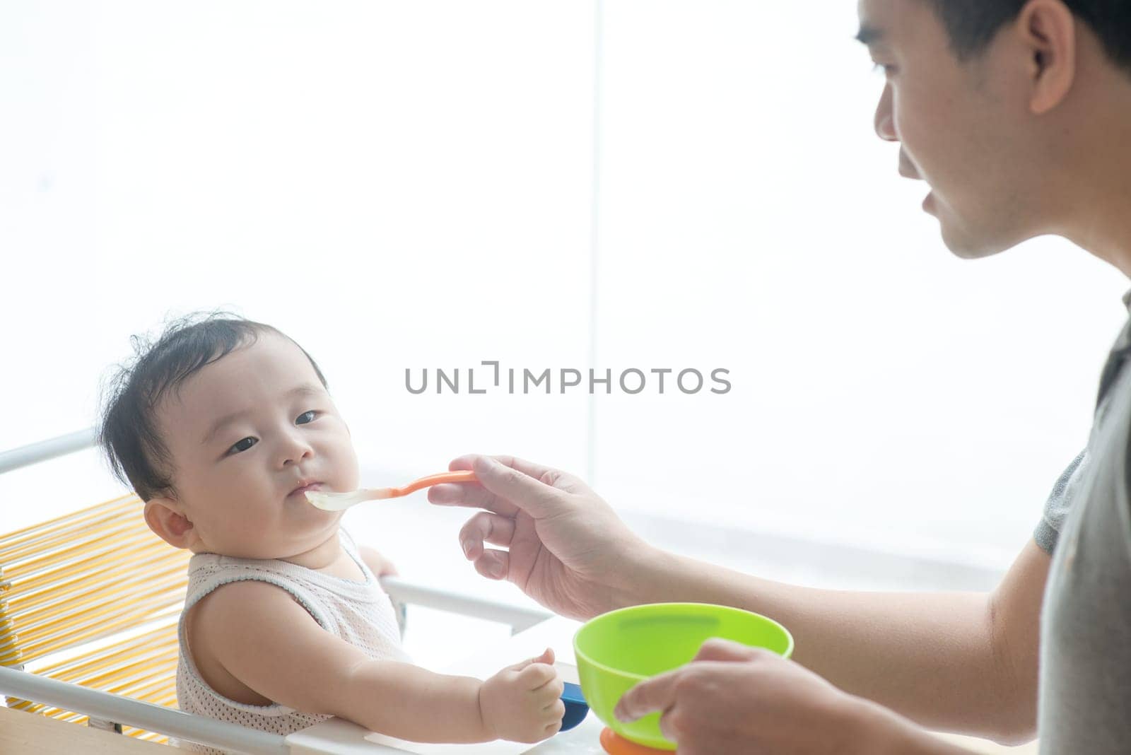 Happy Asian family at home. Father feeding solid food to his 9 months old baby in the kitchen, living lifestyle indoors.