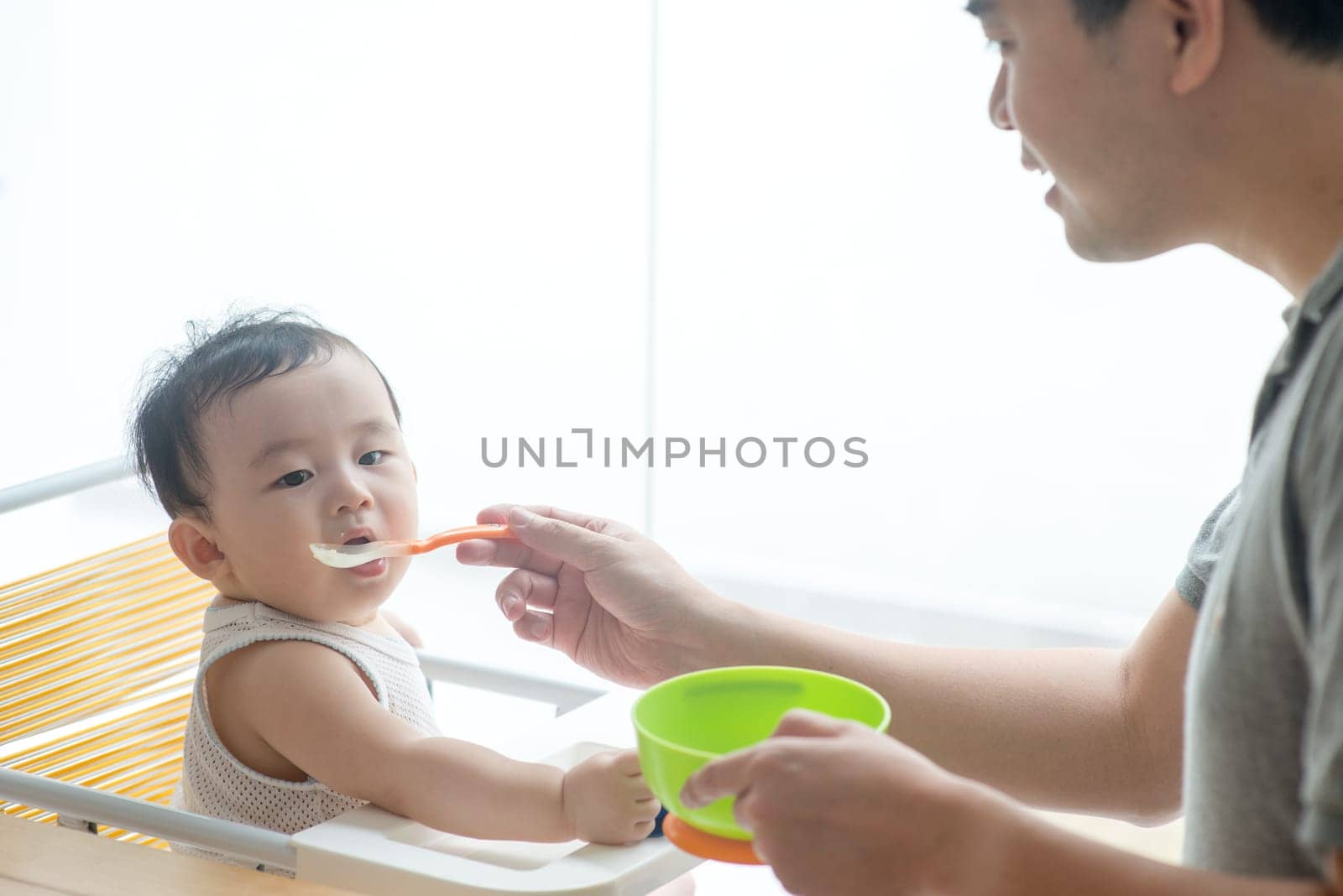 Happy Asian family at home. Father feeding solid food to his 9 months old baby boy in the kitchen, living lifestyle indoors.