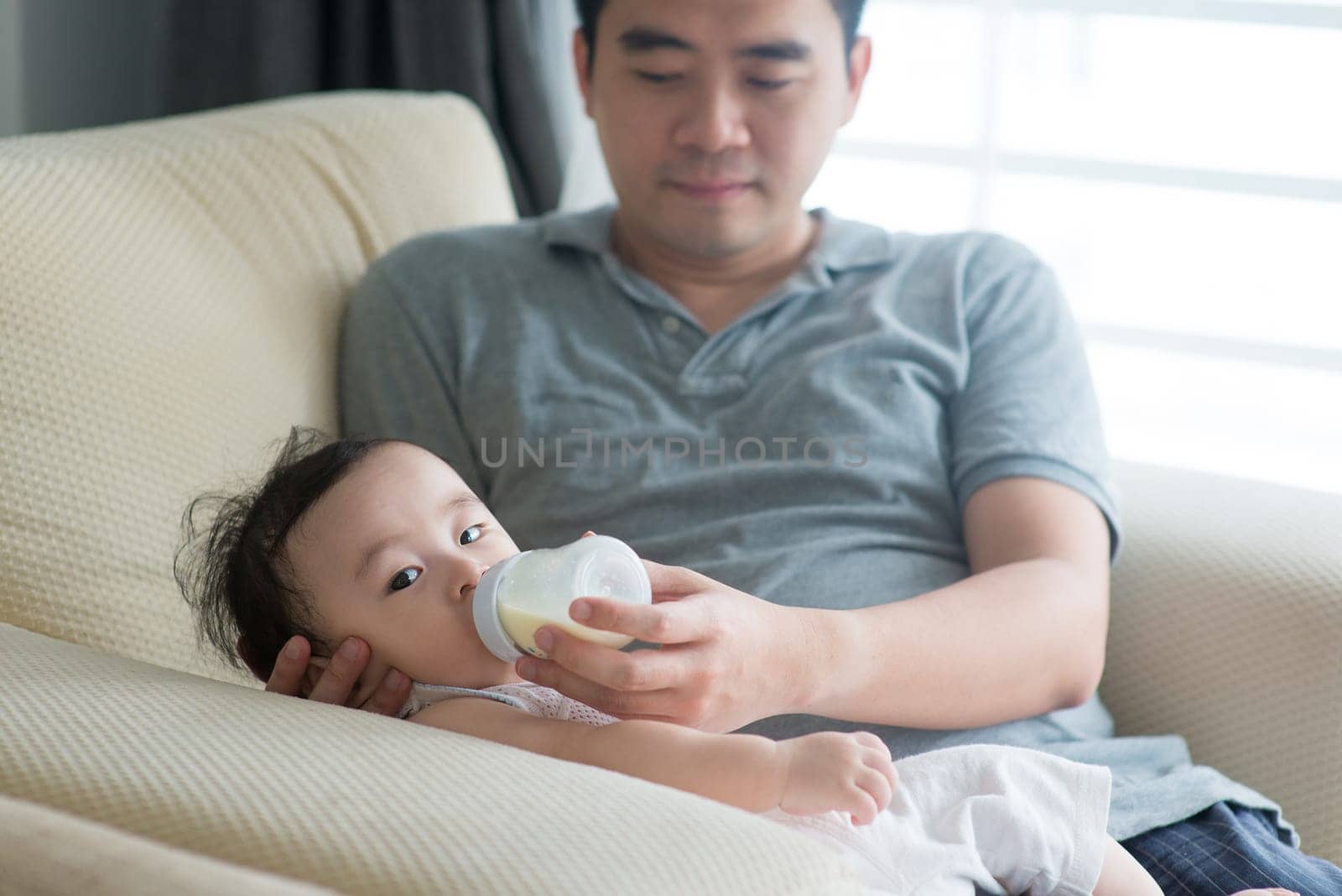 Happy Asian family at home. Father bottle feed formula milk to 9 months old baby, living lifestyle indoors.