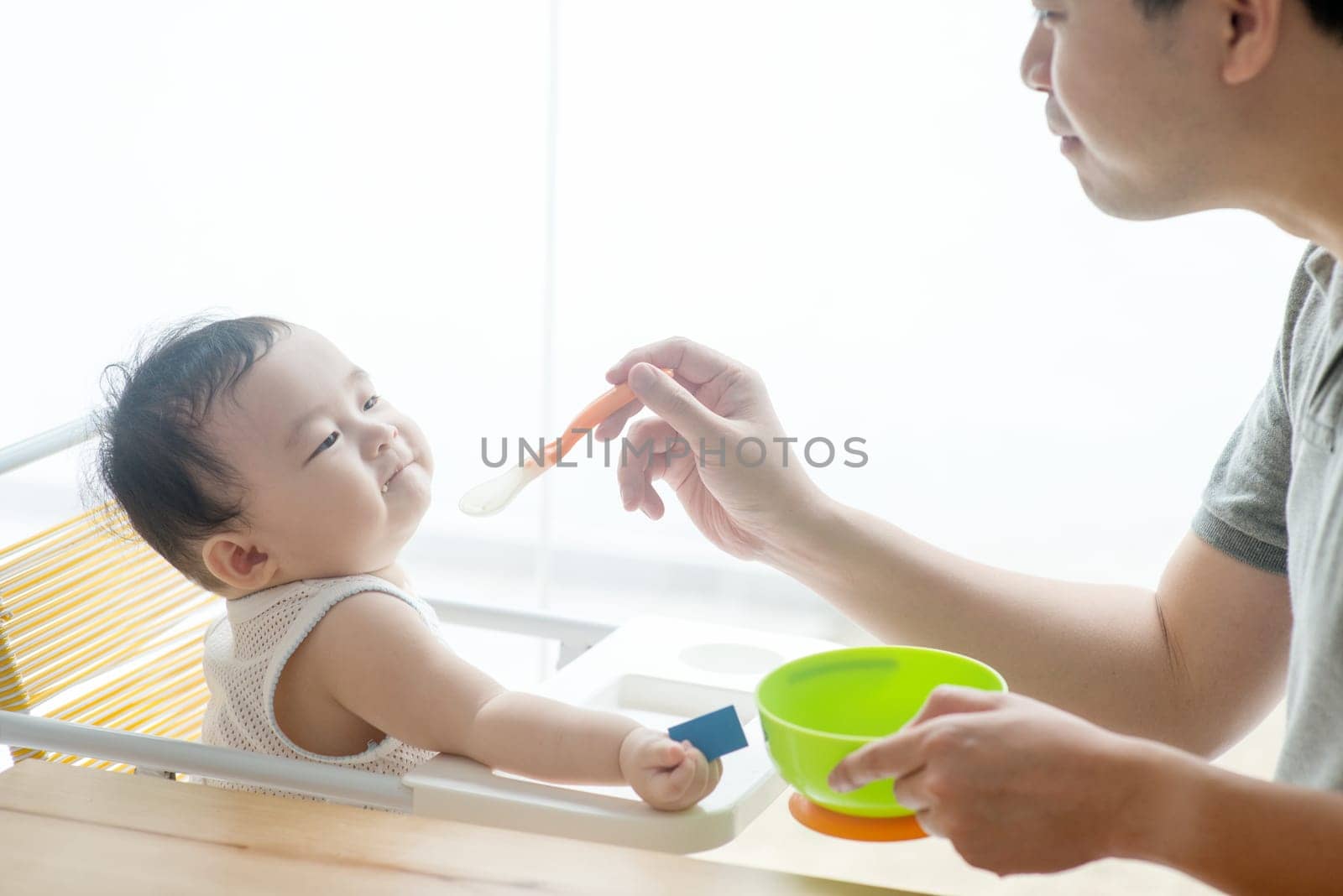 Happy Asian family at home. Father feeding solid food to his 9 months old son in the kitchen, living lifestyle indoors.