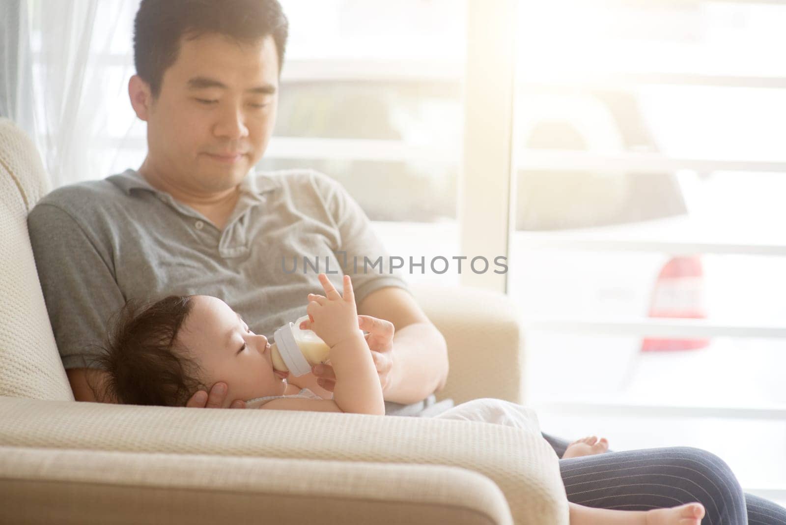 Happy Asian family at home. Father bottle feed formula milk to 9 months old baby boy, living lifestyle indoors.