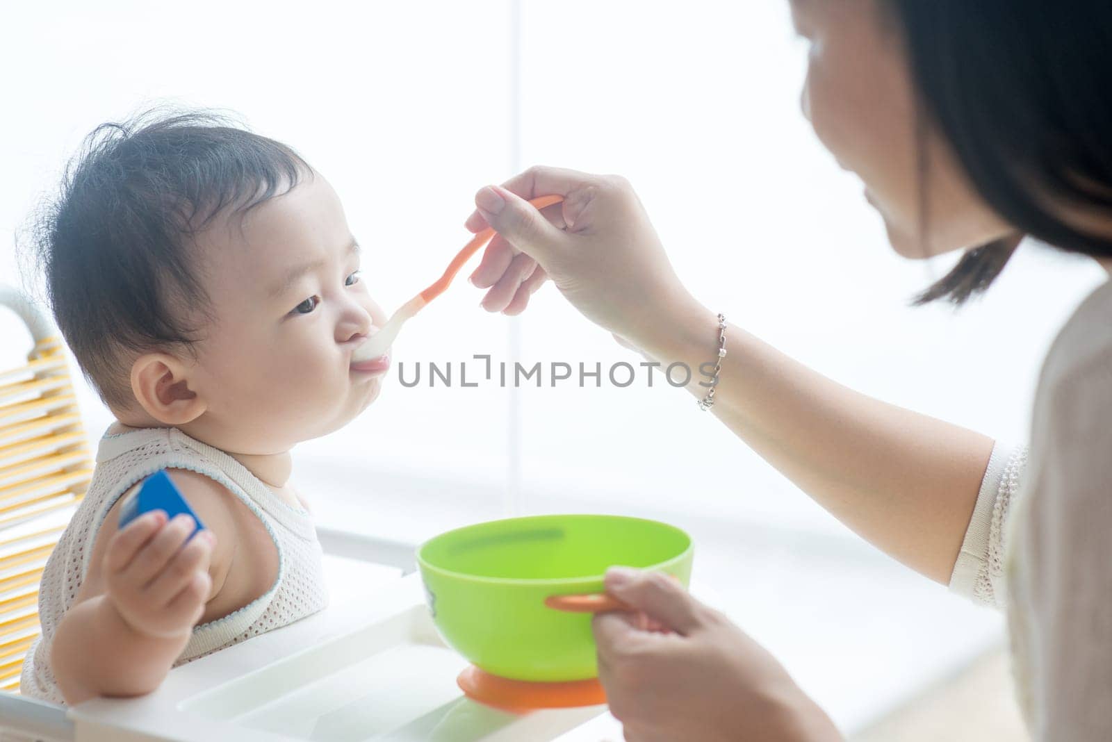 Happy Asian family at home. Mother feeding solid food to 9 months old baby boy in the kitchen, living lifestyle indoors.