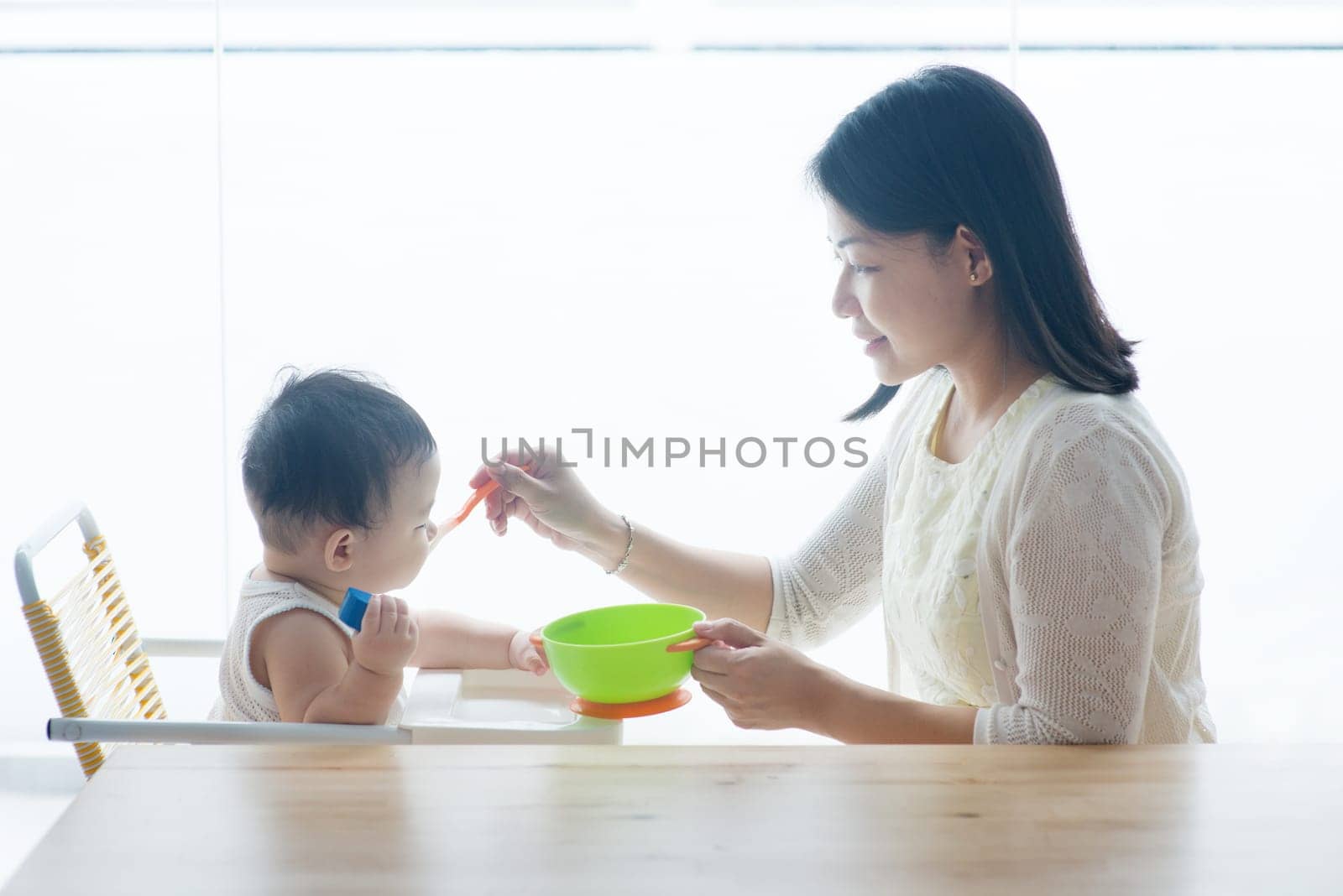 Happy Asian family at home. Mother feeding solid food to 9 months old toddler in the kitchen, living lifestyle indoors.