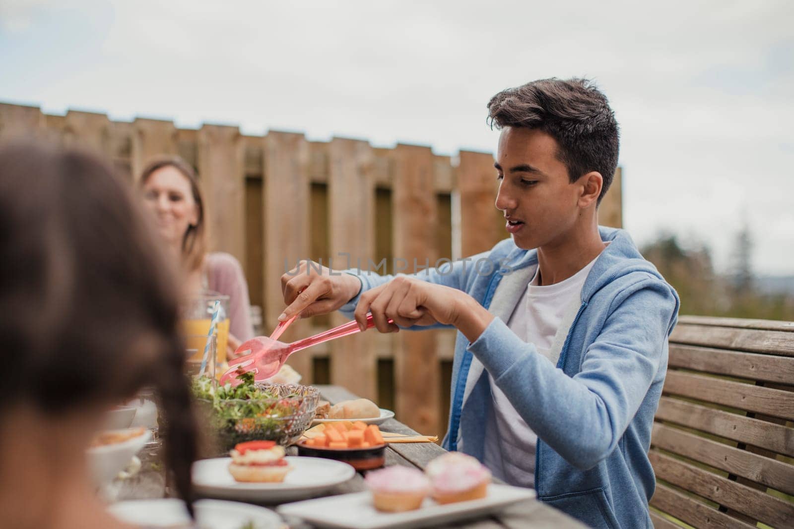 Sitting teenage boy mixing salad at the table outside at brunch.