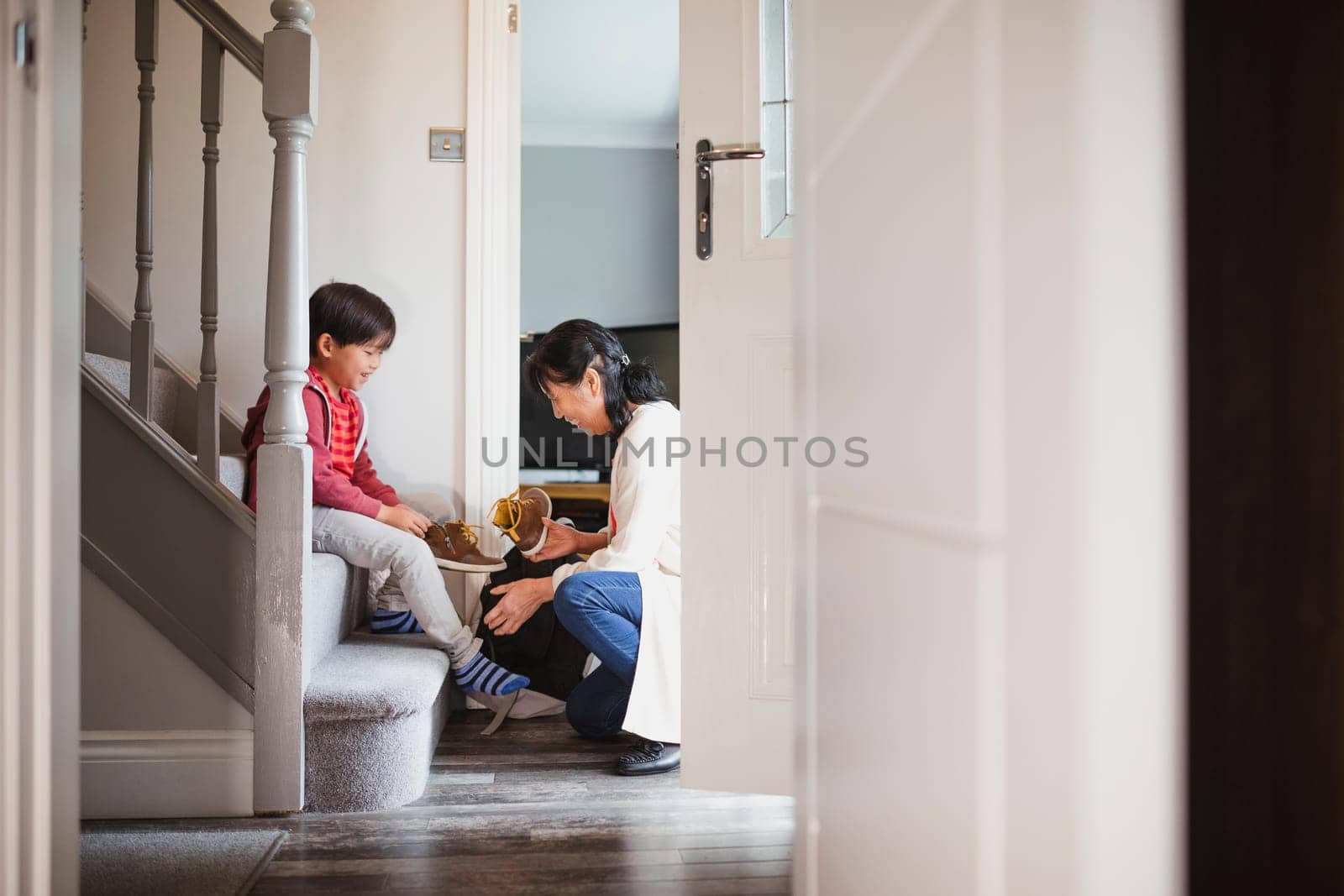 Little boy is sitting at the bottom of the stairs in his home to put his shoes on with help from his grandmother.