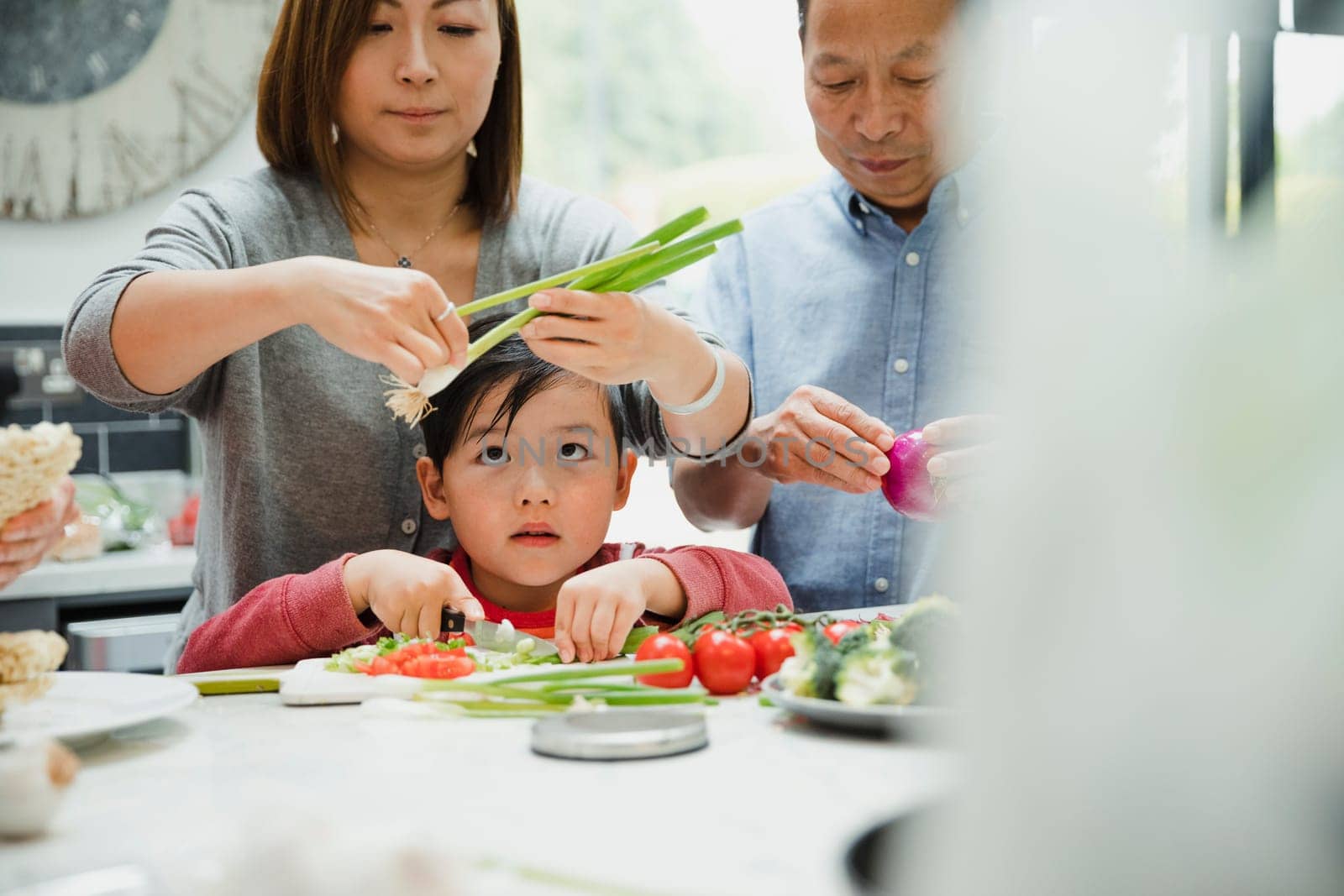 Little boy is learning how to prepare vegetables with his family who are making a stir fry. He is chopping spring onions and tomatoes.