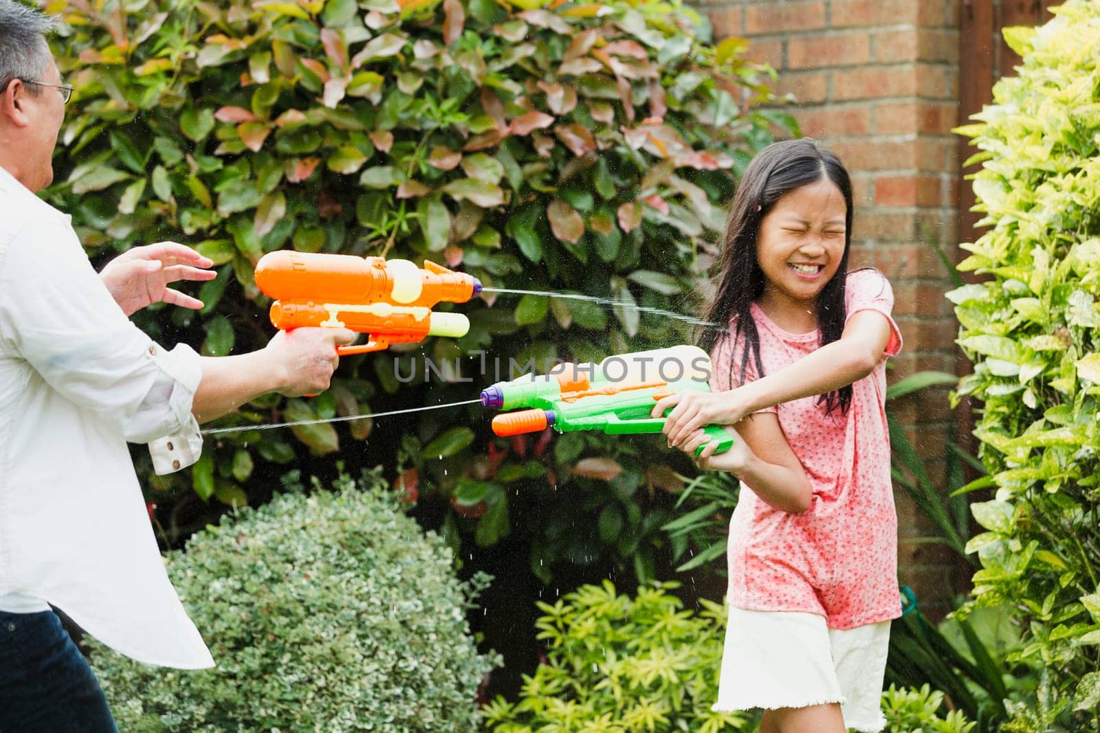 Little girl and her father are having a water fight in the garden with water pistols. The little girl is getting soaked and has her eyes closed as she tries to shoot back.