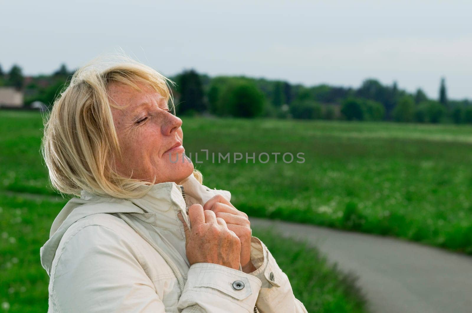 Side outdoor head-and-shoulder view of a blonde mature woman on a green meadow her hands on the lapel and closed eyes breathing deeply.