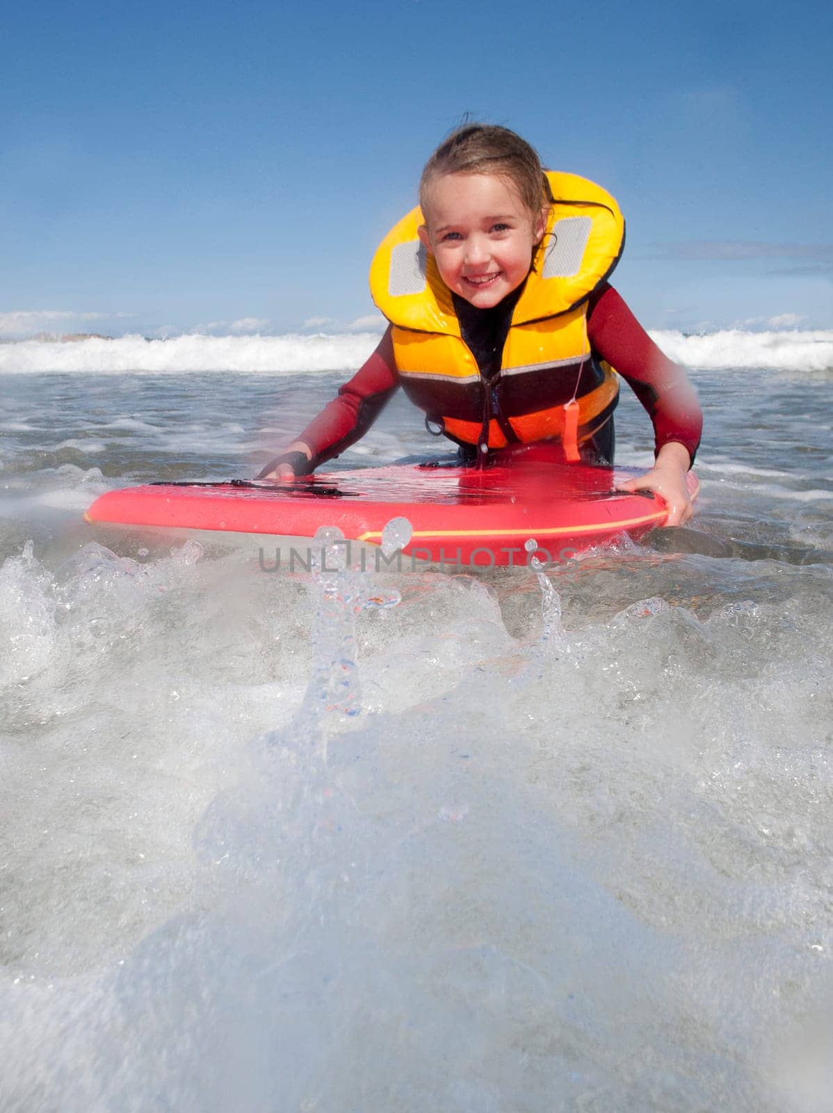 Little girl is smiling for the camera as she bodyboards in the sea with a life jacket on.