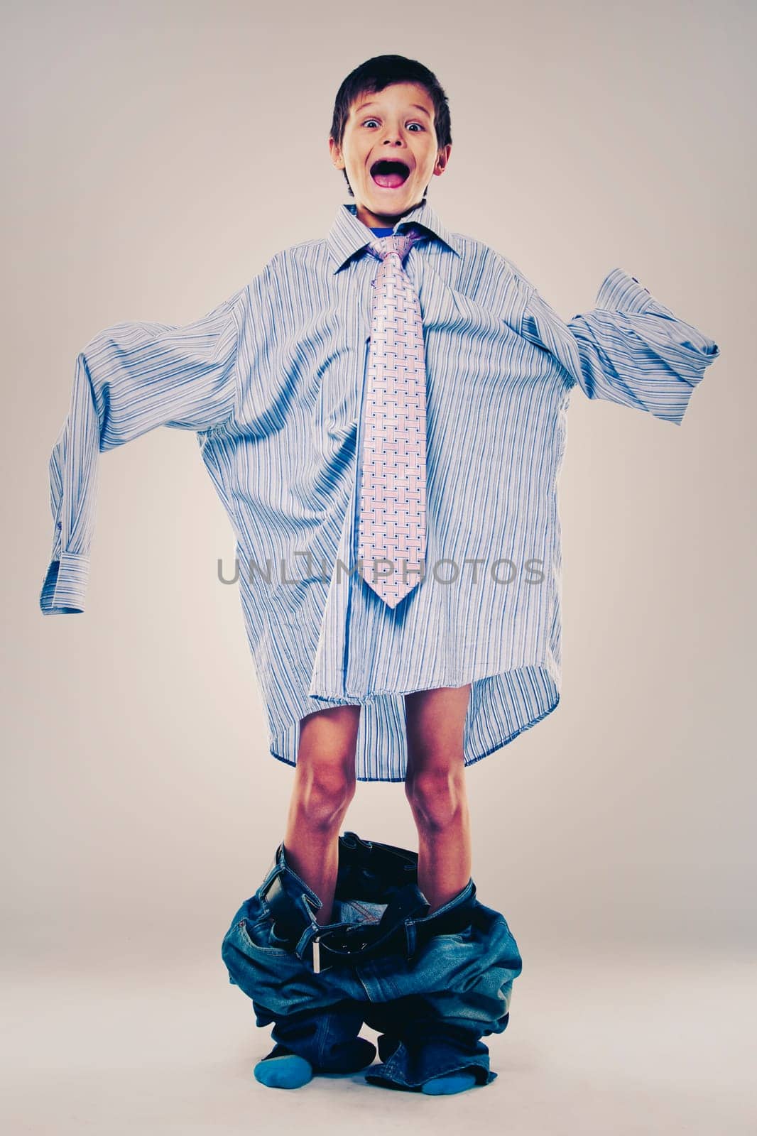 Caucasian boy wearing his Dad's shirt, jeans and tie on light background. He is wearing big adult size clothes which are too big for him.