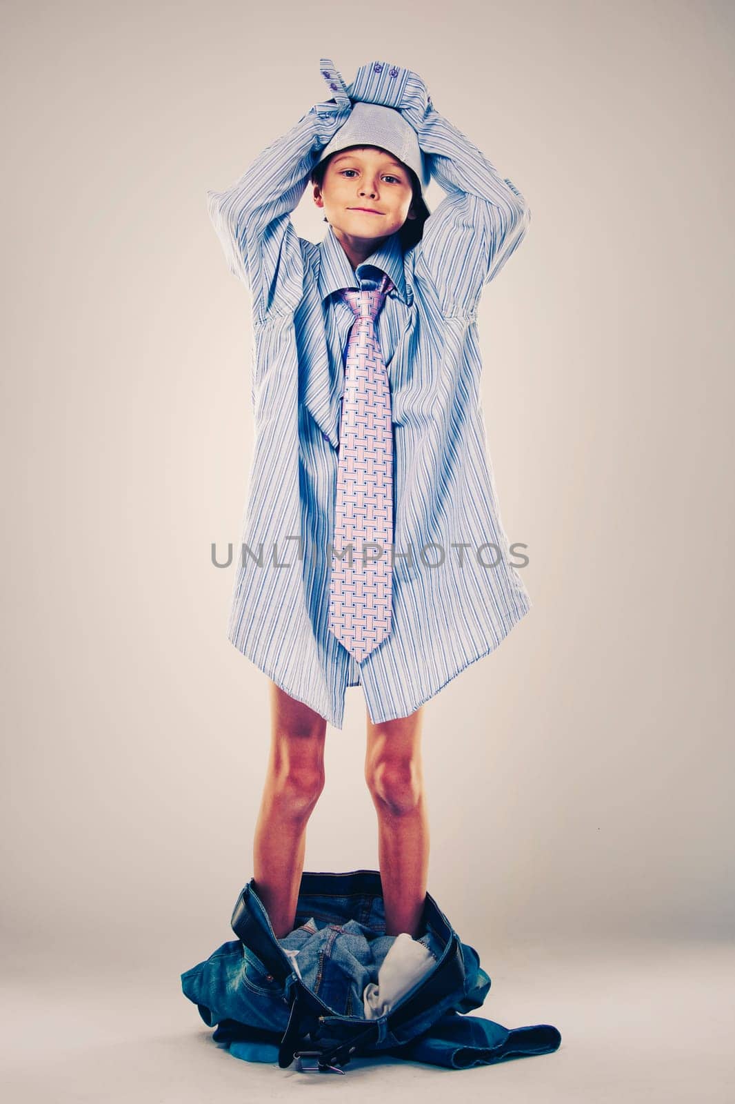 Caucasian boy wearing his Dad's shirt, jeans and tie on light background. He is wearing big adult size clothes which are too big for him.