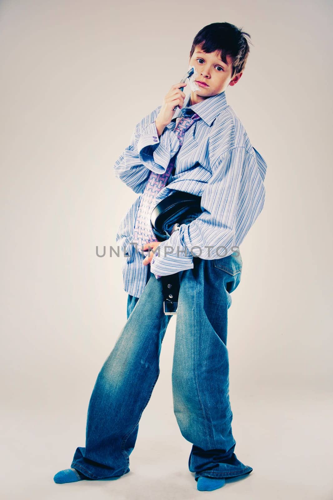 Caucasian boy wearing his Dad's shirt, jeans and tie on light background. He is wearing big adult size clothes which are too big for him.
