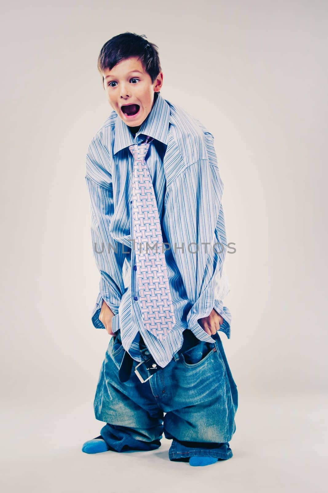 Caucasian boy wearing his Dad's shirt, jeans and tie on light background. He is wearing big adult size clothes which are too big for him.